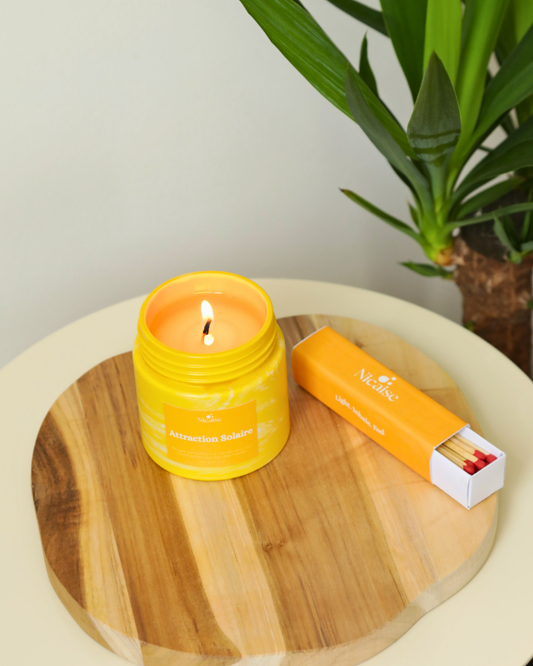 Bougie parfumée Nicaise « Attraction Solaire » allumée en pot jaune marbré, avec boîte d’allumettes longues Nicaise sur planche en bois, plante verte en fond.