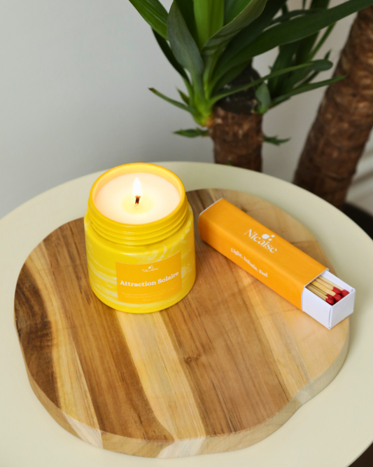 Bougie parfumée Nicaise « Attraction Solaire » allumée en pot jaune marbré, avec boîte d’allumettes longues Nicaise sur planche en bois.