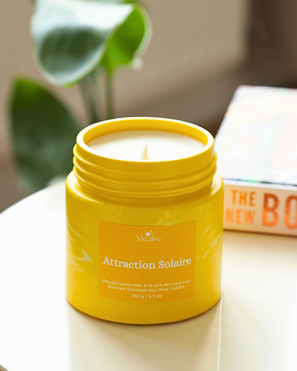 Bougie parfumée Nicaise « Attraction Solaire » en pot jaune marbré, posée sur une table avec plante en arrière-plan.