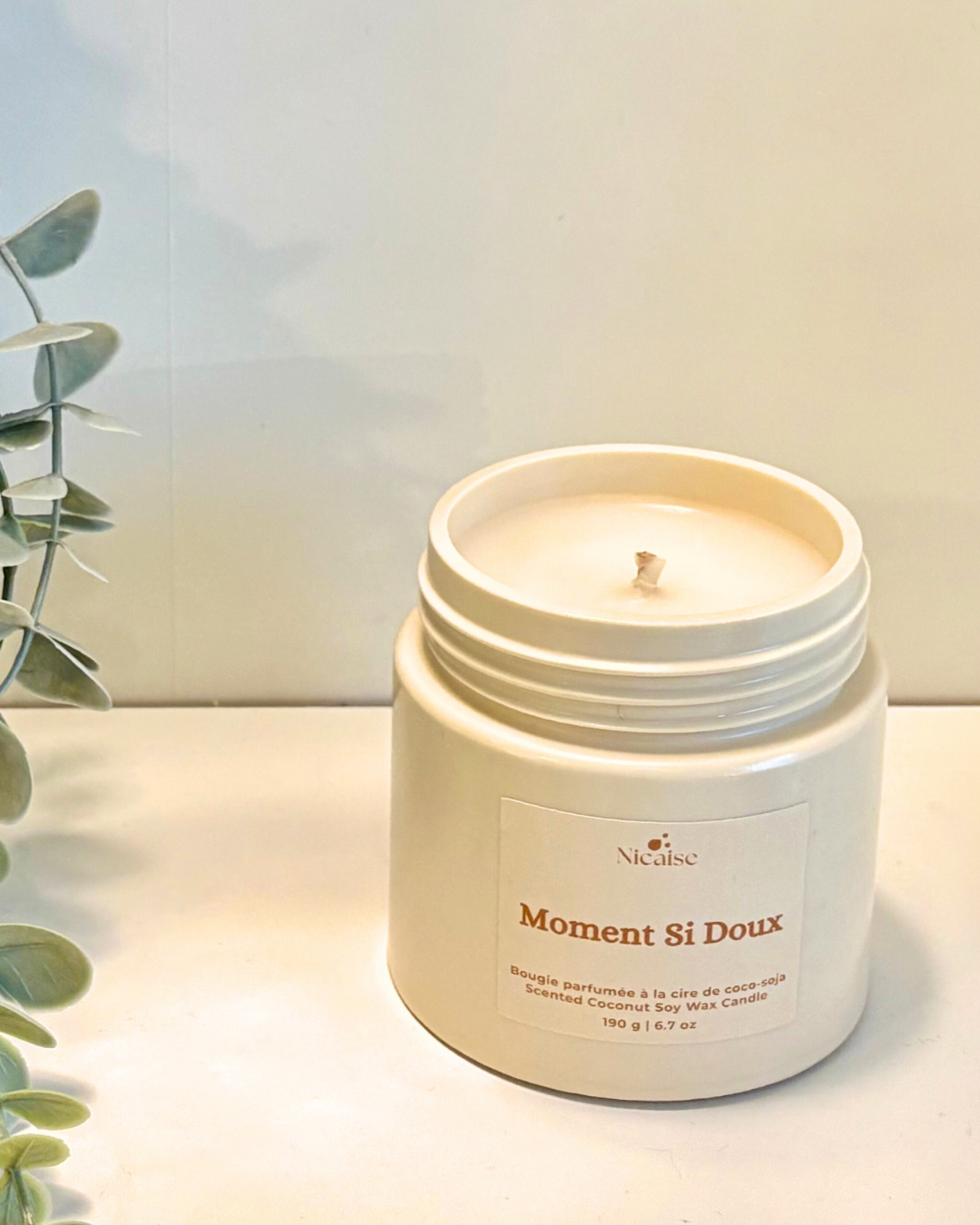 Bougie parfumée Nicaise « Moment Si Doux » en pot blanc sans couvercle, posée sur une étagère avec plante décorative.
