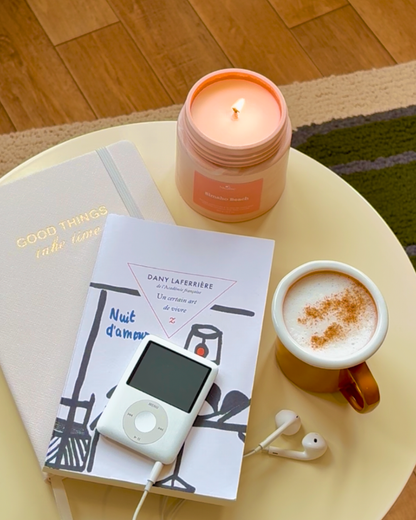 Bougie parfumée Simaho Beach allumée sur une table basse en métal écru, décoration d'intérieur douce et cosy, bloc notes, livres, mug de chai latte.