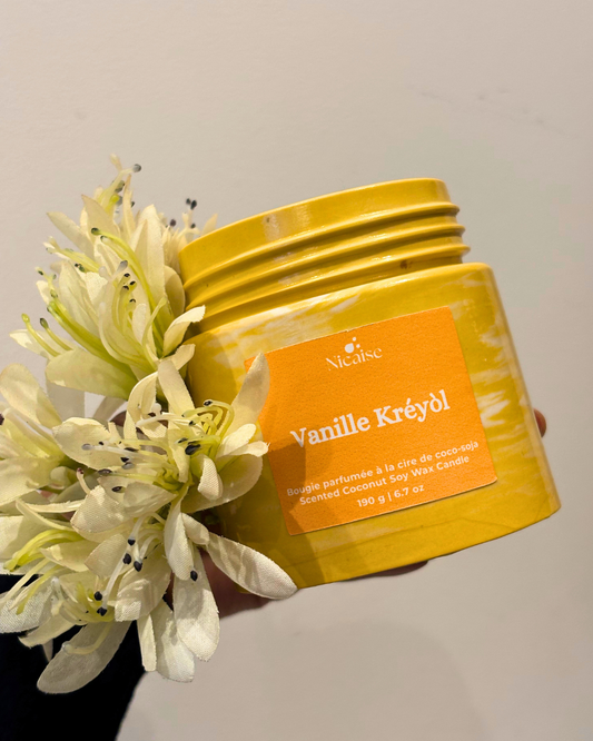 Bougie parfumée Nicaise « Vanille Kréyòl » en pot jaune marbré, tenue à la main avec fleurs blanches.