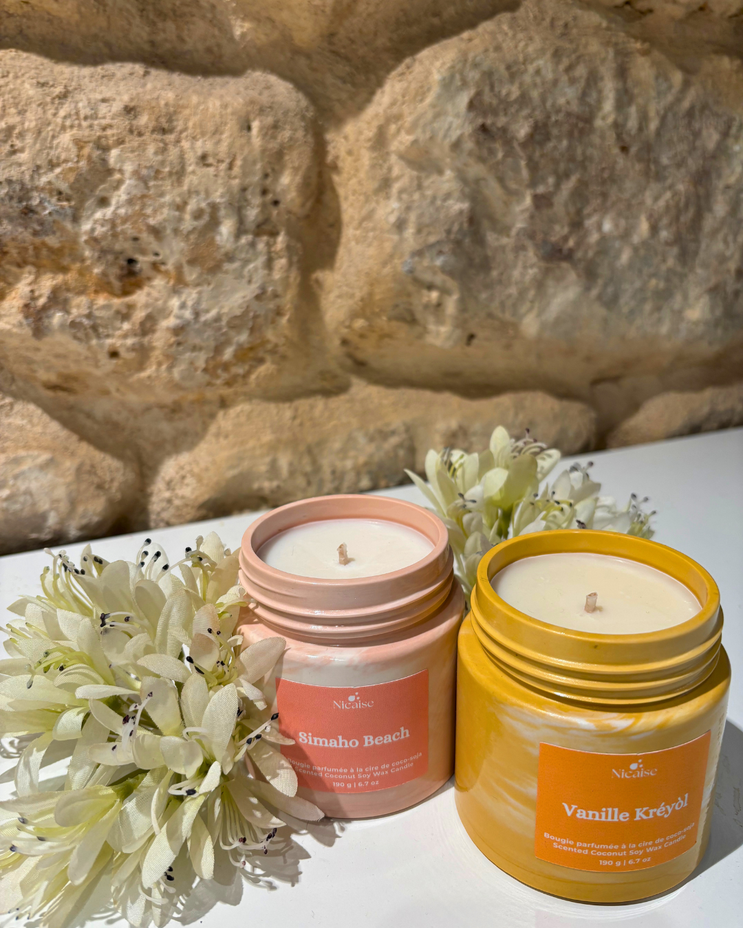 Deux bougies parfumées artisanales Nicaise en pots marbrés, entourées de lys blanc : Simaho Beach et Vanille Kréyòl.
