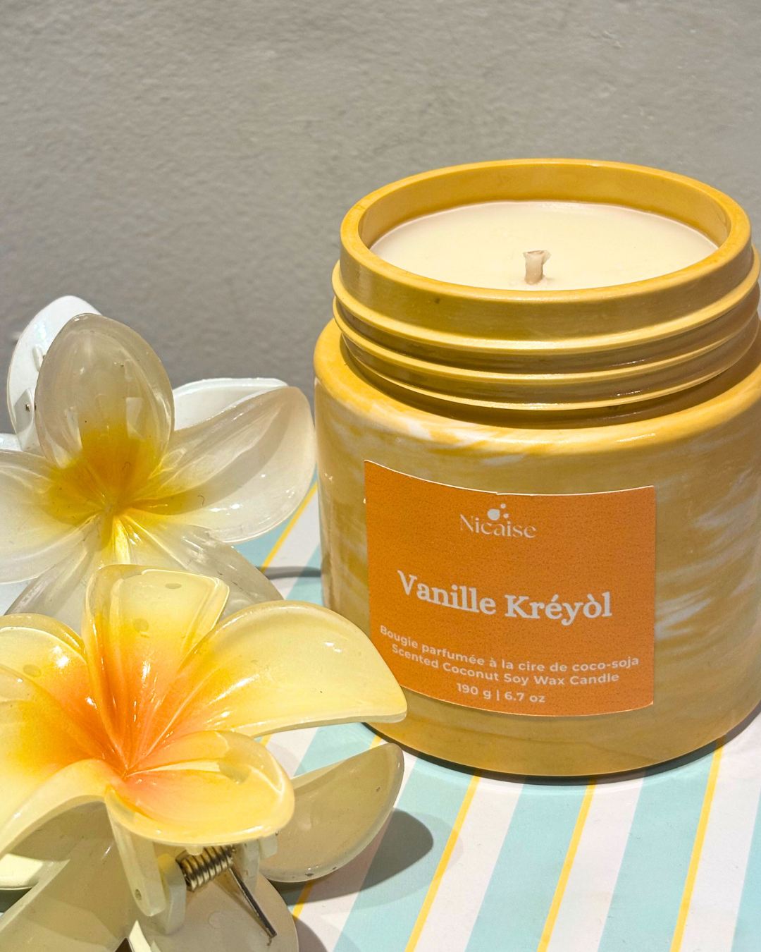 Bougie parfumée Nicaise « Vanille Kréyòl » en pot jaune marbré, posée près de fleurs décoratives sur fond rayé.