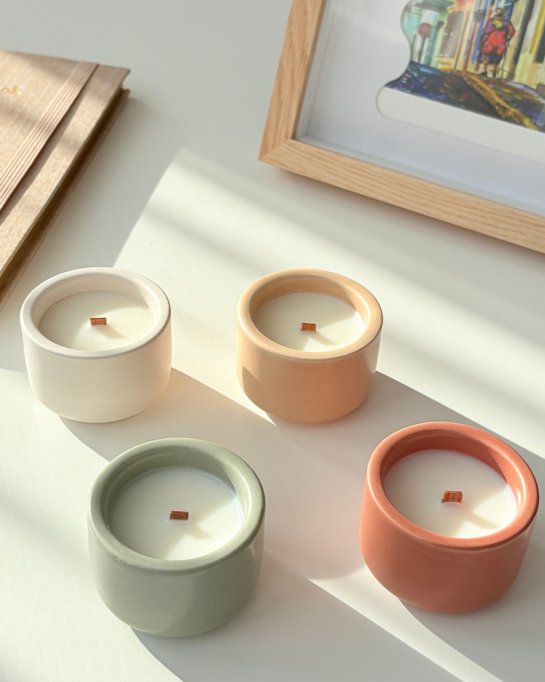 Quatre bougies parfumées Nicaise en pots colorés (blanc, beige, vert sauge, terracotta) avec mèches en bois, posées sur une table lumineuse.