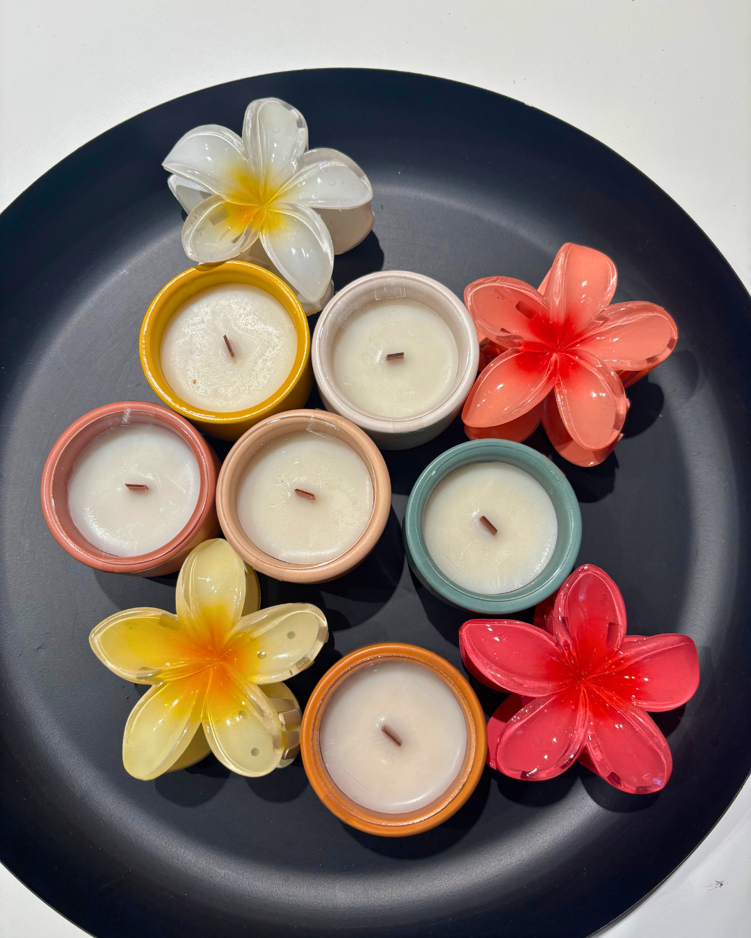 Six mini bougies parfumées Nicaise en pots colorés avec mèches en bois, présentées sur un plateau noir avec fleurs décoratives.