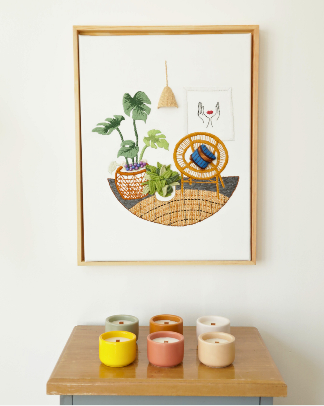 Six mini bougies parfumées Nicaise en pots colorés avec mèches en bois, présentées sur un meuble en bois vernis sous un tableau brodé en relief avec un cadre en bois.